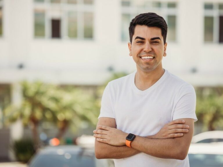 Vereador Cryslan, de São José, que está denunciando o uso de recursos do governo federal para financiar invasões em Santa Catarina, sorri de camiseta branca e braços cruzados. Paisagem urbana ao fundo.