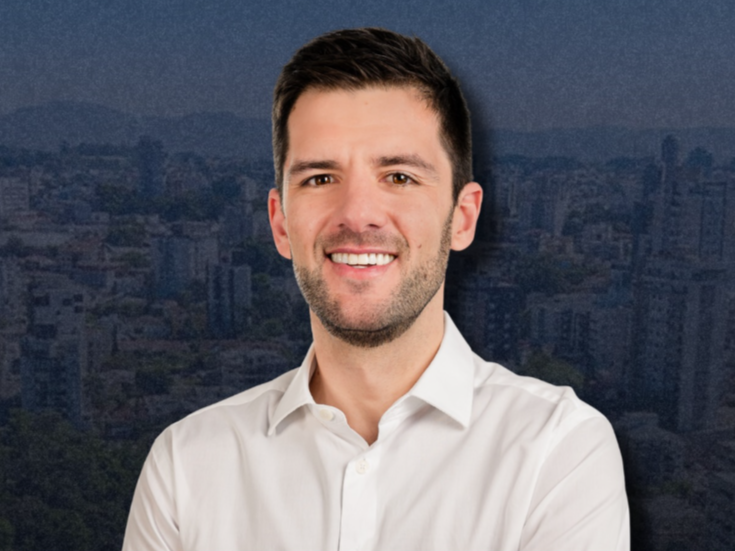 Guilherme Cé, mandatário do Partido Novo e novo prefeito de Lajeado, sorri e ao fundo dele aparece a imagem do centro da cidade com um filtro azul escuro.